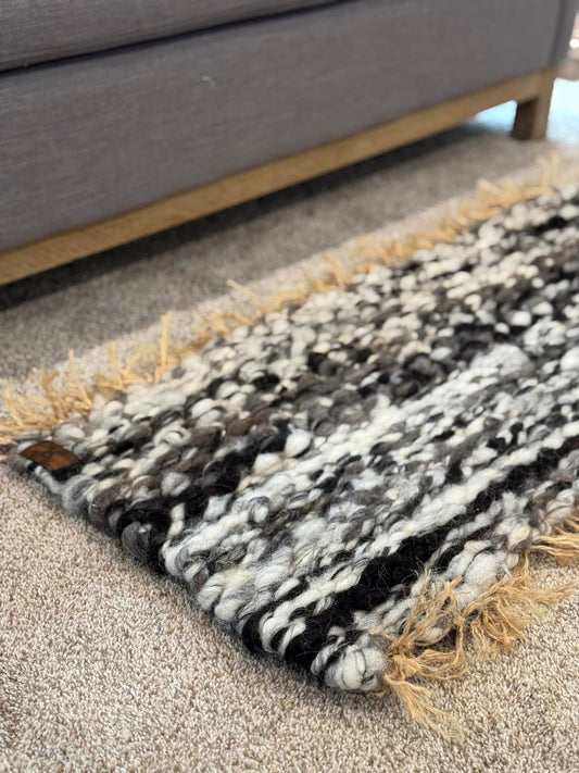"A close-up of a rustic, handwoven grey and white wool rug displayed on a carpeted floor next to a grey chair at Wild Daisy Homestead."
